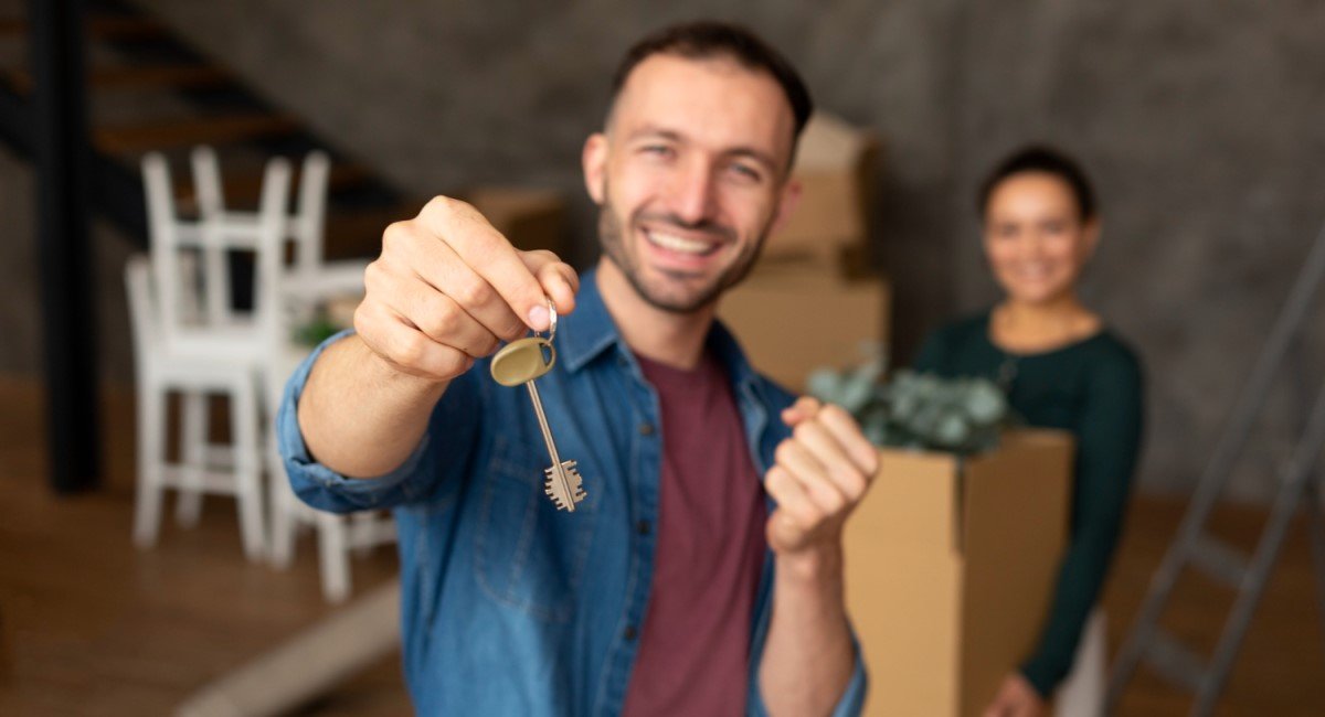 Homem sorridente segurando uma chave de casa, com uma mulher desfocada ao fundo carregando uma caixa de mudança.