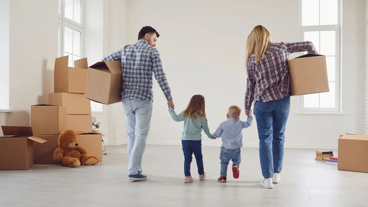 Uma família de quatro pessoas entra em uma sala ampla e vazia carregando caixas de mudança. Eles estão de costas, caminhando em direção a uma janela iluminada.