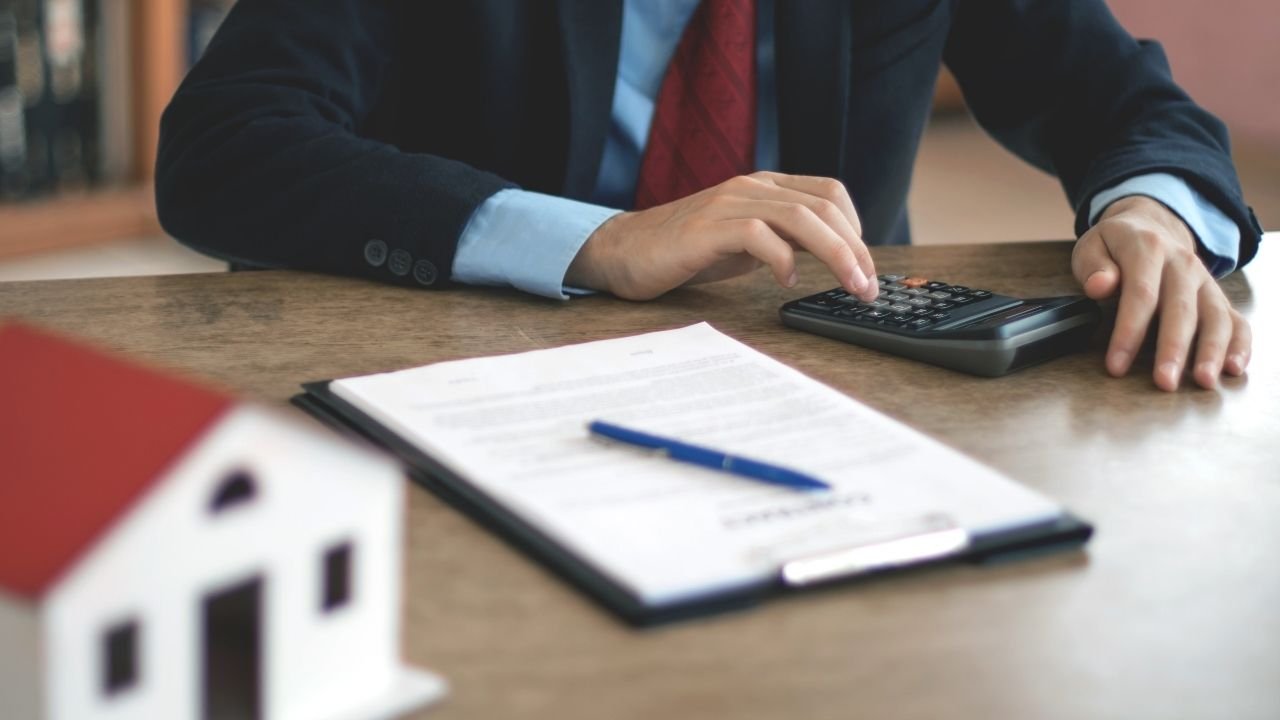 profissional usando calculadora sobre mesa com contrato de compra e venda de imóvel e miniatura de casa desfocada.