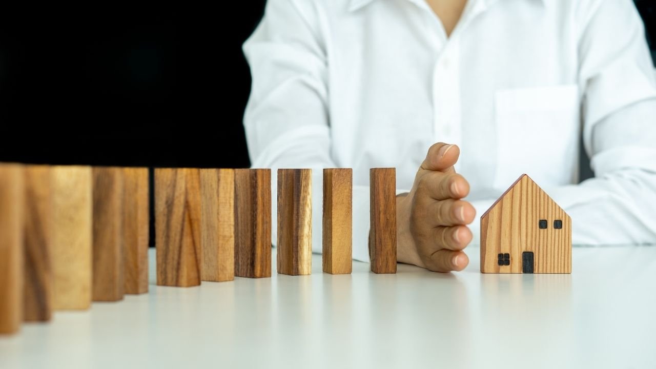 Uma pessoa usa a mão para proteger uma miniatura de casa de blocos de madeira caindo, ilustrando a ideia de proteção. A imagem representa como o seguro fiança serve como uma barreira, protegendo o imóvel e as partes envolvidas de riscos financeiros.