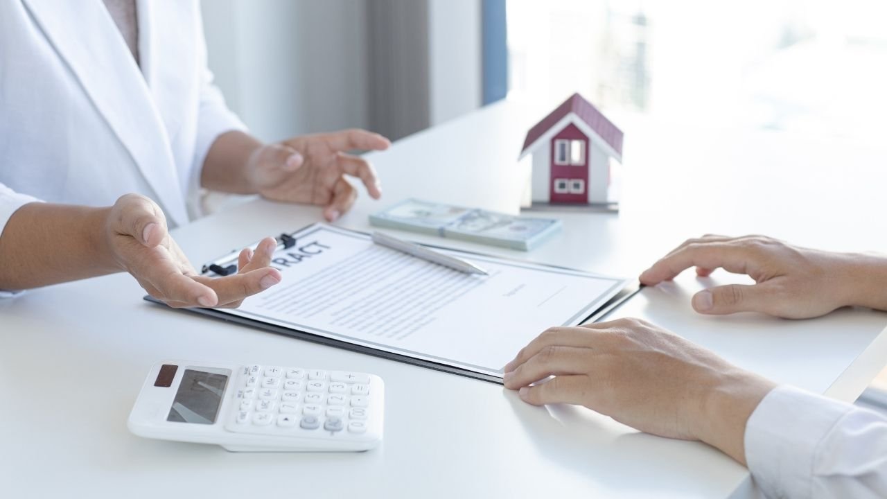 Duas pessoas conversam em uma mesa com um contrato de locação, uma calculadora e uma miniatura de casa. A imagem demonstra a discussão e o planejamento financeiro ao considerar o seguro fiança como alternativa para alugar um imóvel.