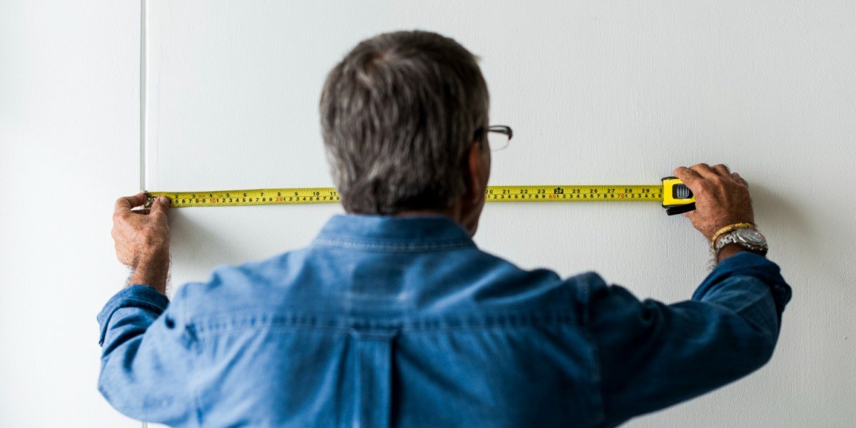 Homem de camisa jeans azul, medindo a parede com uma fita métrica amarela