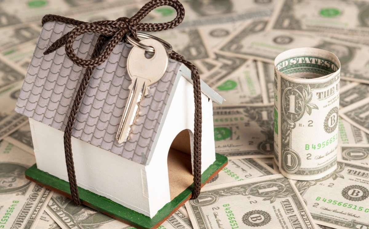 Modelo de casa em miniatura amarrada com uma chave, rodeada por notas de dólar, simbolizando financiamento imobiliário e investimentos em propriedades.