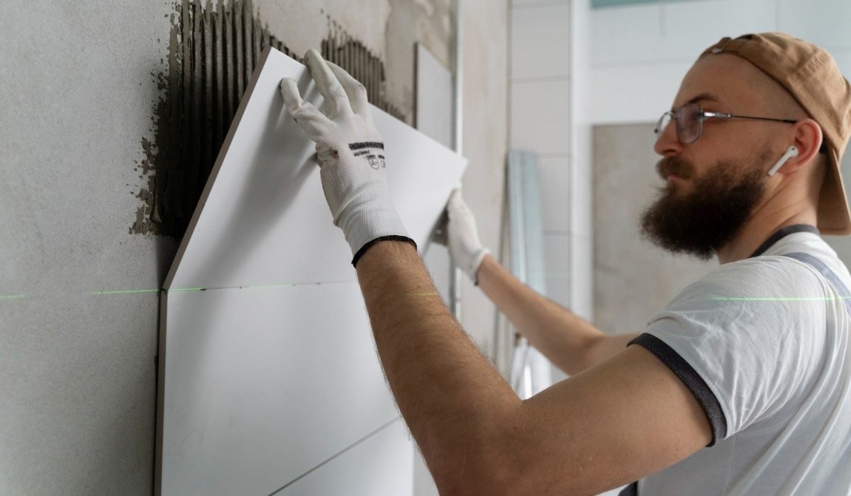 Trabalhador instalando azulejos em uma parede. A imagem mostra um homem de barba e boné ao contrário, usando luvas brancas, aplicando um grande azulejo branco em uma parede cinza. O trabalhador está em um ambiente interno, provavelmente um banheiro ou cozinha, e está focado no processo de reforma. A cena enfatiza a instalação de revestimentos e a melhoria de interiores.

