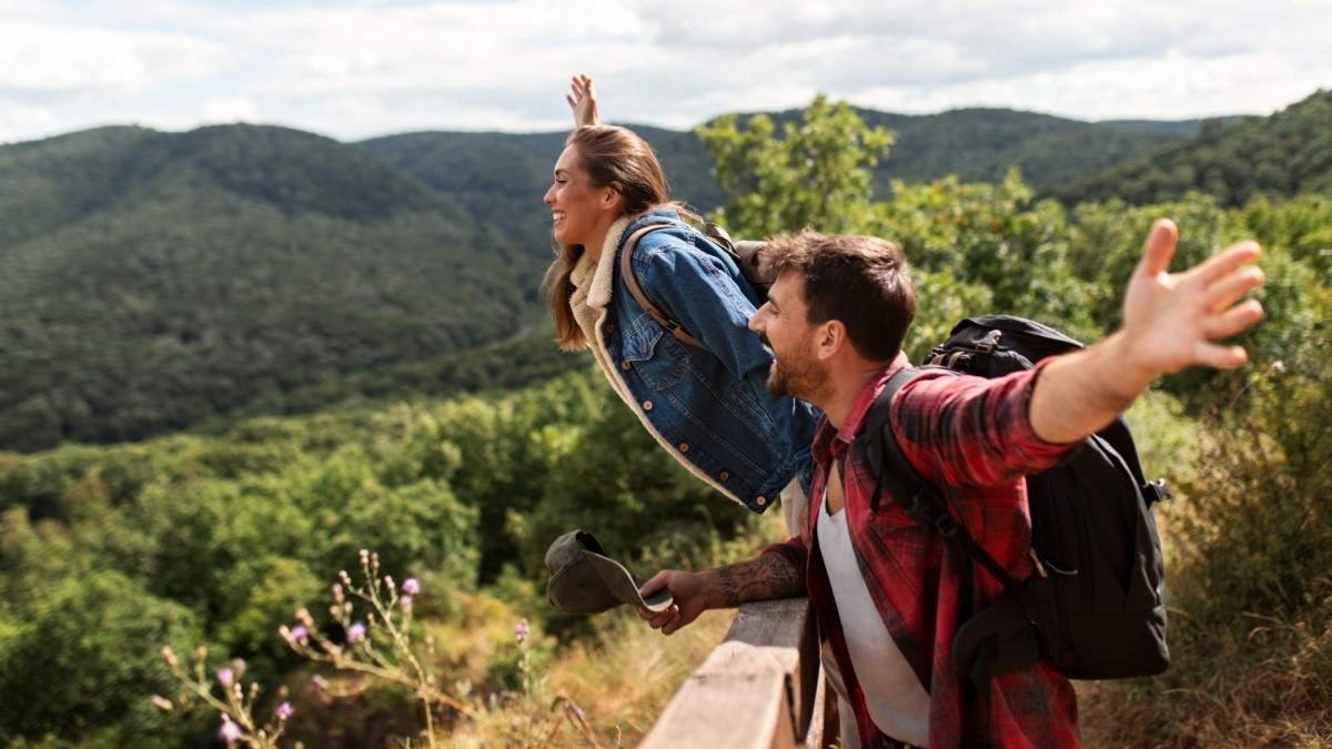 Dois amigos felizes em uma trilha, de braços abertos, em um ambiente montanhoso e verdejante