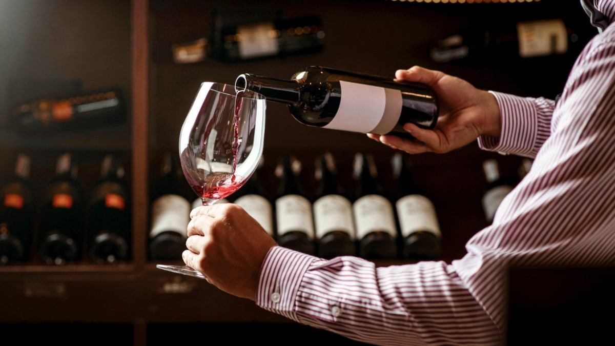essoa segurando uma garrafa de vinho e servindo vinho tinto em uma taça, com várias garrafas de vinho dispostas em uma prateleira ao fundo