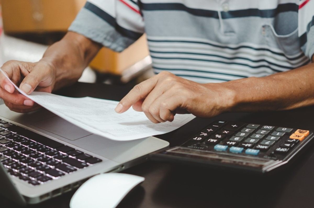 Pessoa revisando documentos financeiros com um laptop e uma calculadora em uma mesa, simbolizando a análise de despesas e orçamento pessoal.