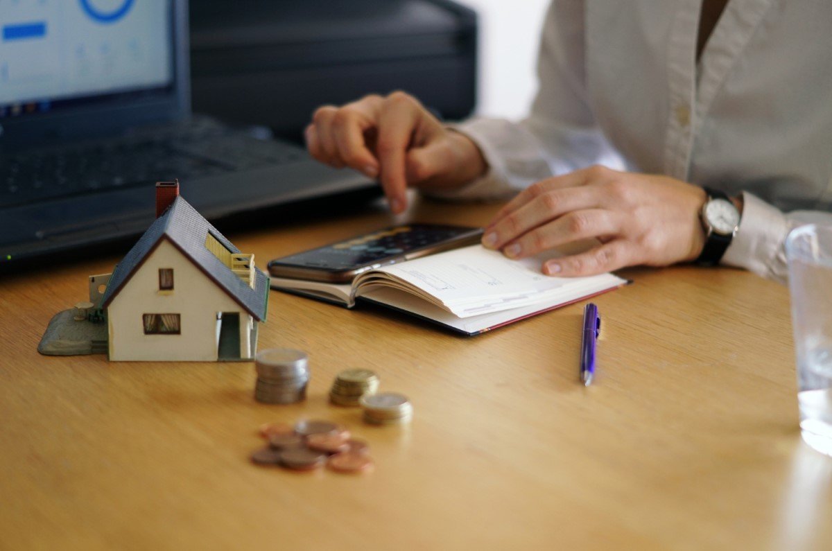 Pessoa fazendo anotações em um caderno com uma calculadora e um laptop na mesa, ao lado de um modelo de casa e moedas, simbolizando planejamento financeiro e investimentos imobiliários.

