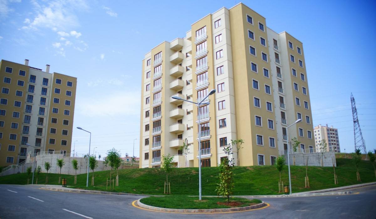 Prédio residencial de múltiplos andares, com fachada bege e detalhes brancos, cercado por uma área verde e ruas asfaltadas.