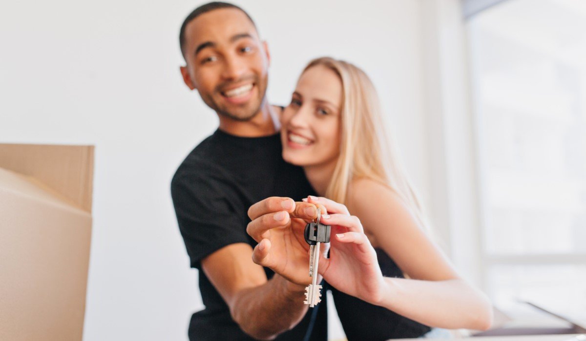 Casal sorridente segurando a chave de sua nova casa, com caixas de mudança ao fundo