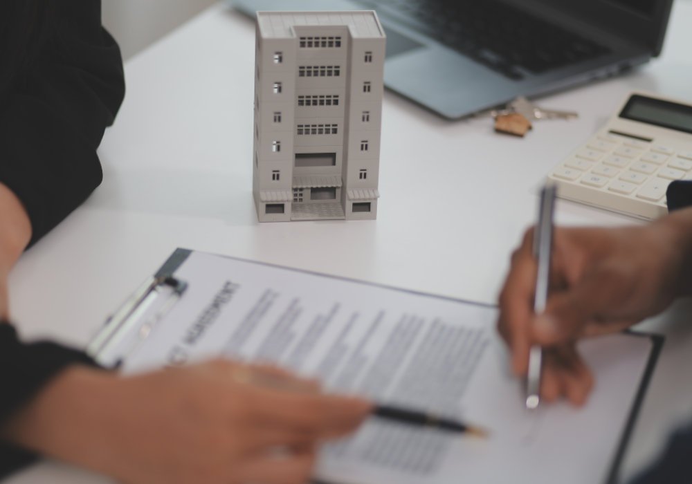 Pessoa assinando um contrato de compra de imóvel com uma maquete de prédio residencial ao fundo, em um ambiente de escritório