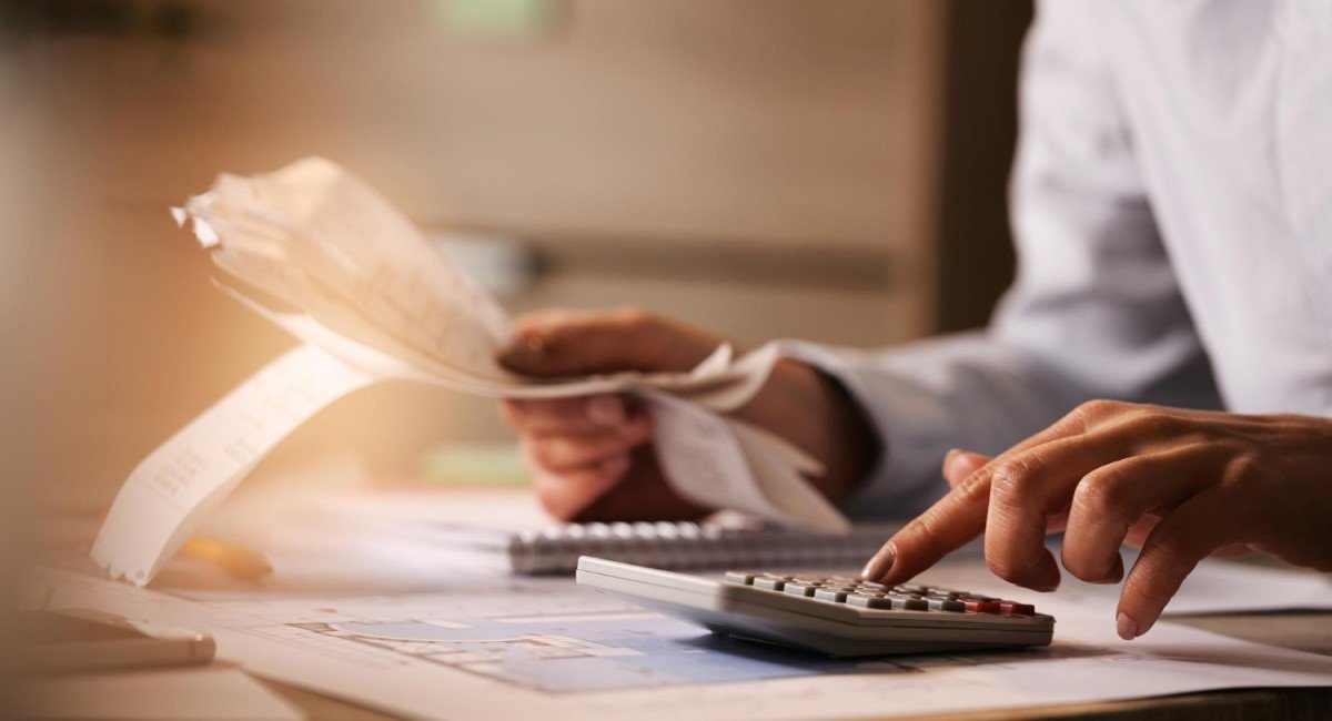  Mulher analisando documentos financeiros e usando uma calculadora em uma mesa com papéis e gráficos espalhados, em um ambiente de escritório iluminado.