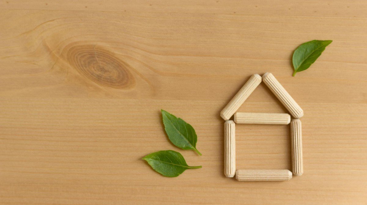 Estrutura de uma casa feita de palitos de madeira com folhas verdes ao lado, representando a construção ecológica e o conceito de lar sustentável, sobre uma superfície de madeira clara.