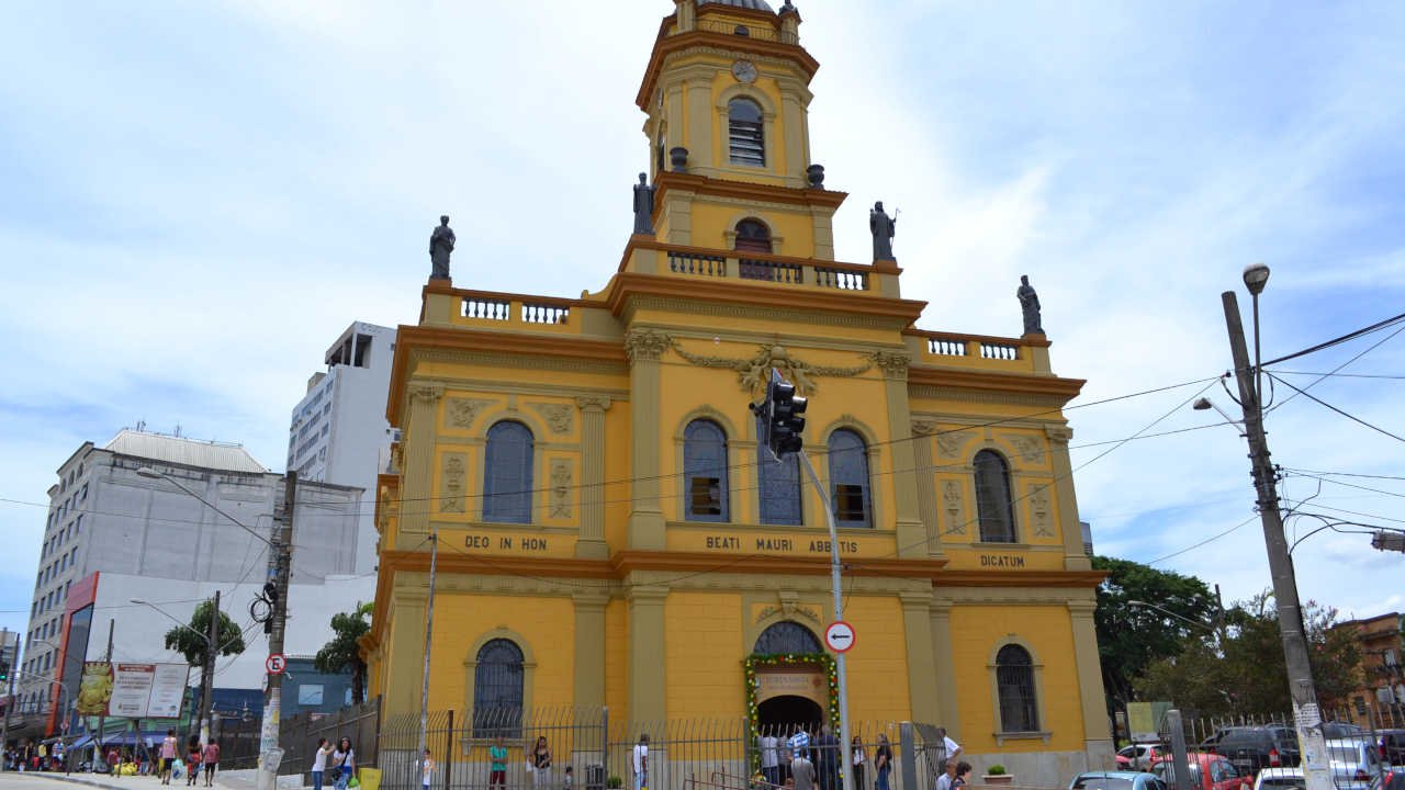 Foto da grande igreja amarela com estátuas no topo e uma alta torre central, cercada por ruas urbanas e prédios.