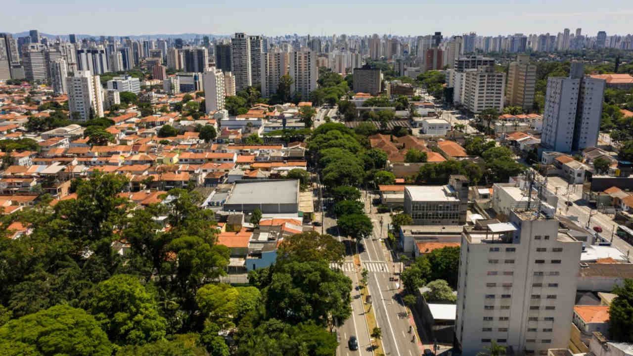 Foto do bairro com muitas casas baixas, grandes árvores e prédios altos ao fundo, capturando uma mistura de verde e cidade.