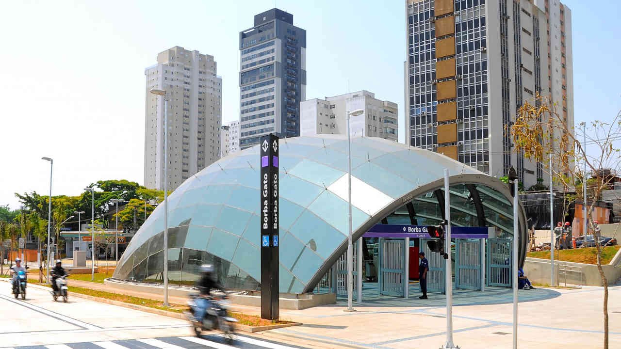 Foto da entrada curva de vidro da estação, com prédios altos próximos e algumas pessoas andando e passando de moto.