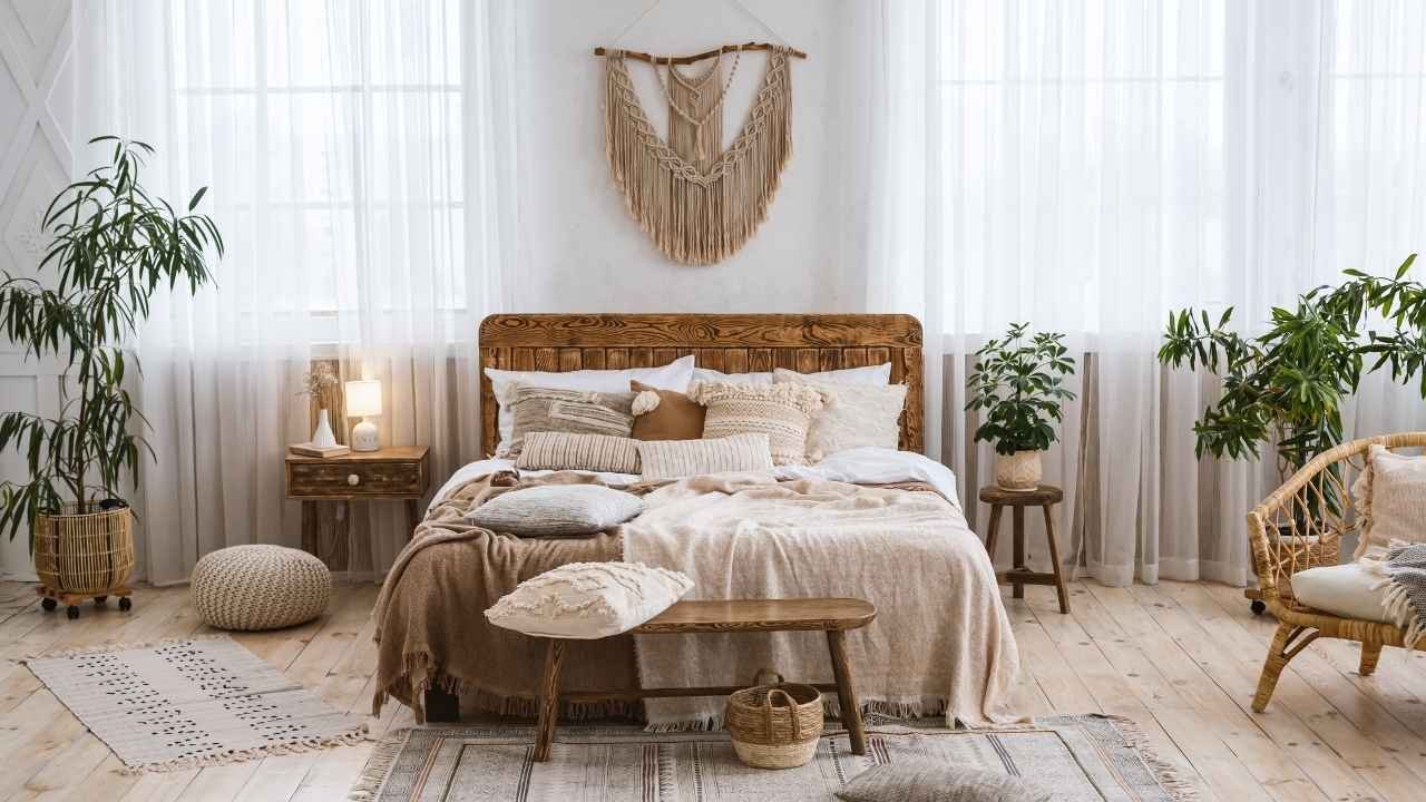 Quarto decorado no estilo boho, com cama de madeira, almofadas e mantas em tons neutros, além de plantas e cortinas translúcidas complementando o ambiente acolhedor.