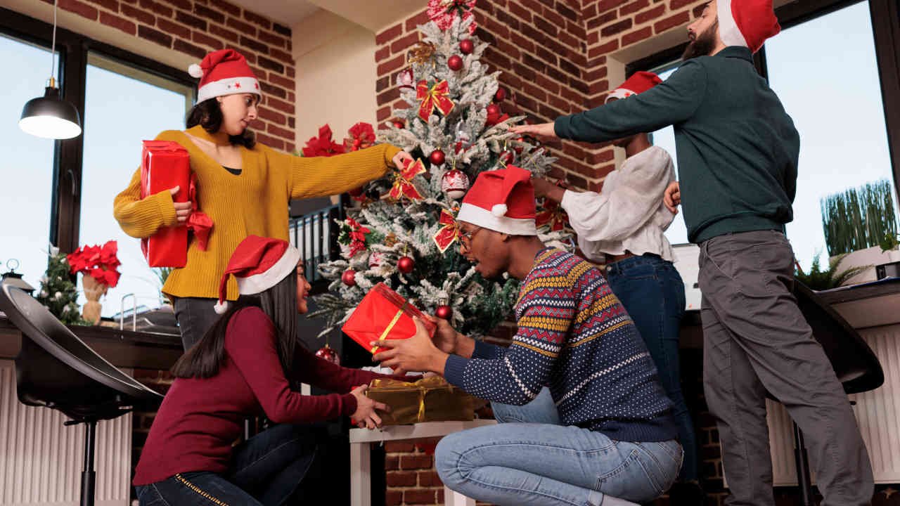 Grupo de amigos com gorros de Natal decorando uma árvore em um ambiente com parede de tijolos aparentes.