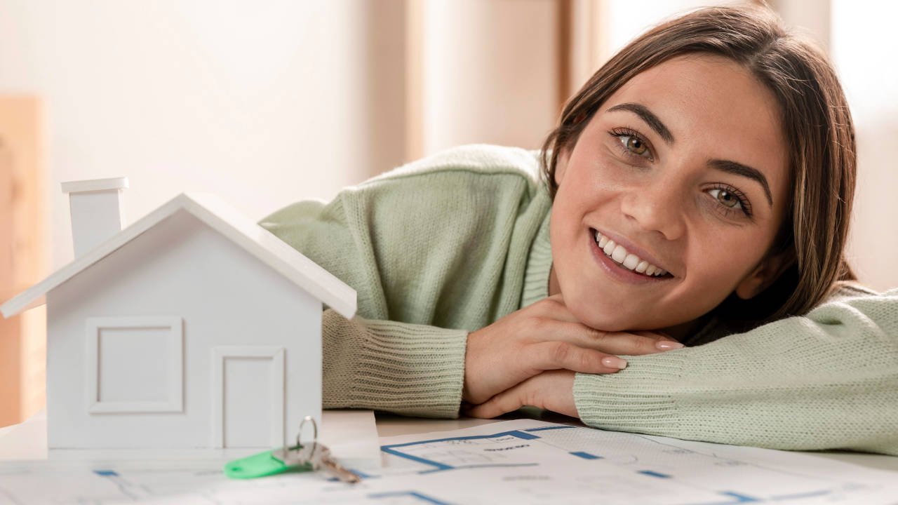 Mulher sorridente com um modelo de casa ao lado, representando a conquista ou o planejamento de um novo lar.