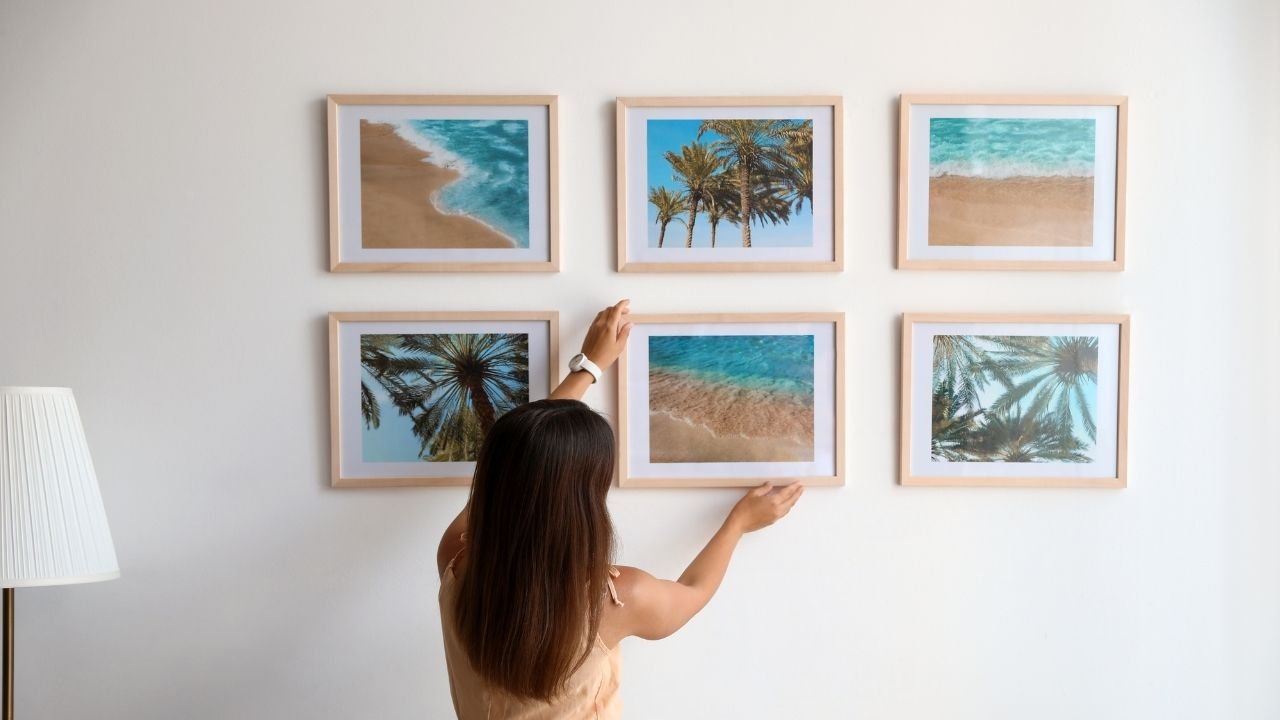 Mulher ajustando uma composição de quadros decorativos com fotografias de paisagens de praia e palmeiras.