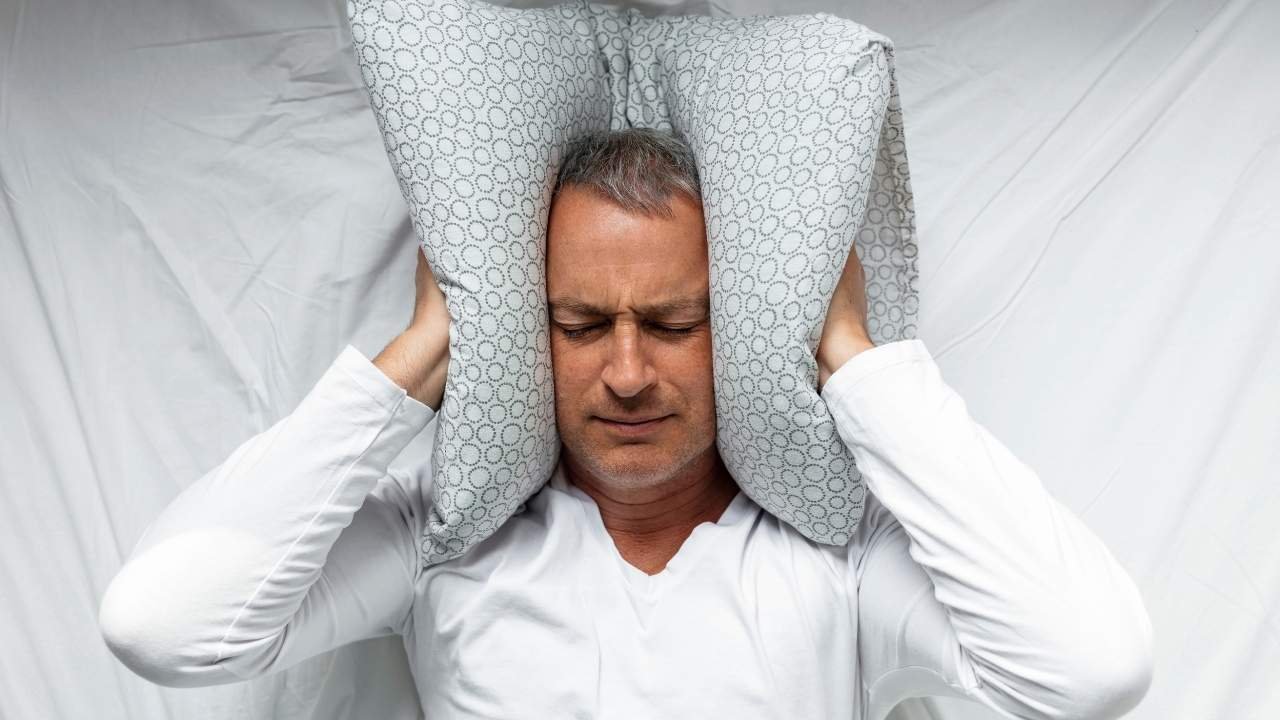 Homem na cama cobrindo os ouvidos com travesseiro.