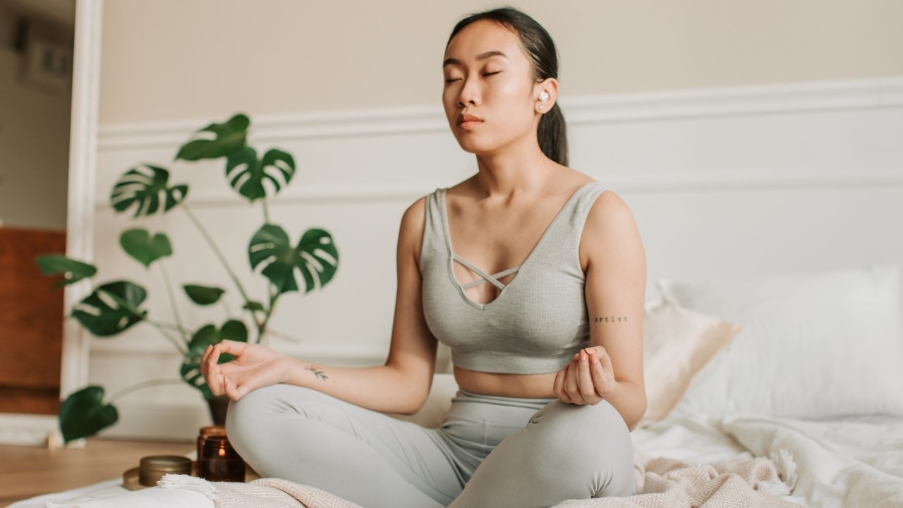 Mulher meditando em ambiente tranquilo com plantas decorativas ao fundo.