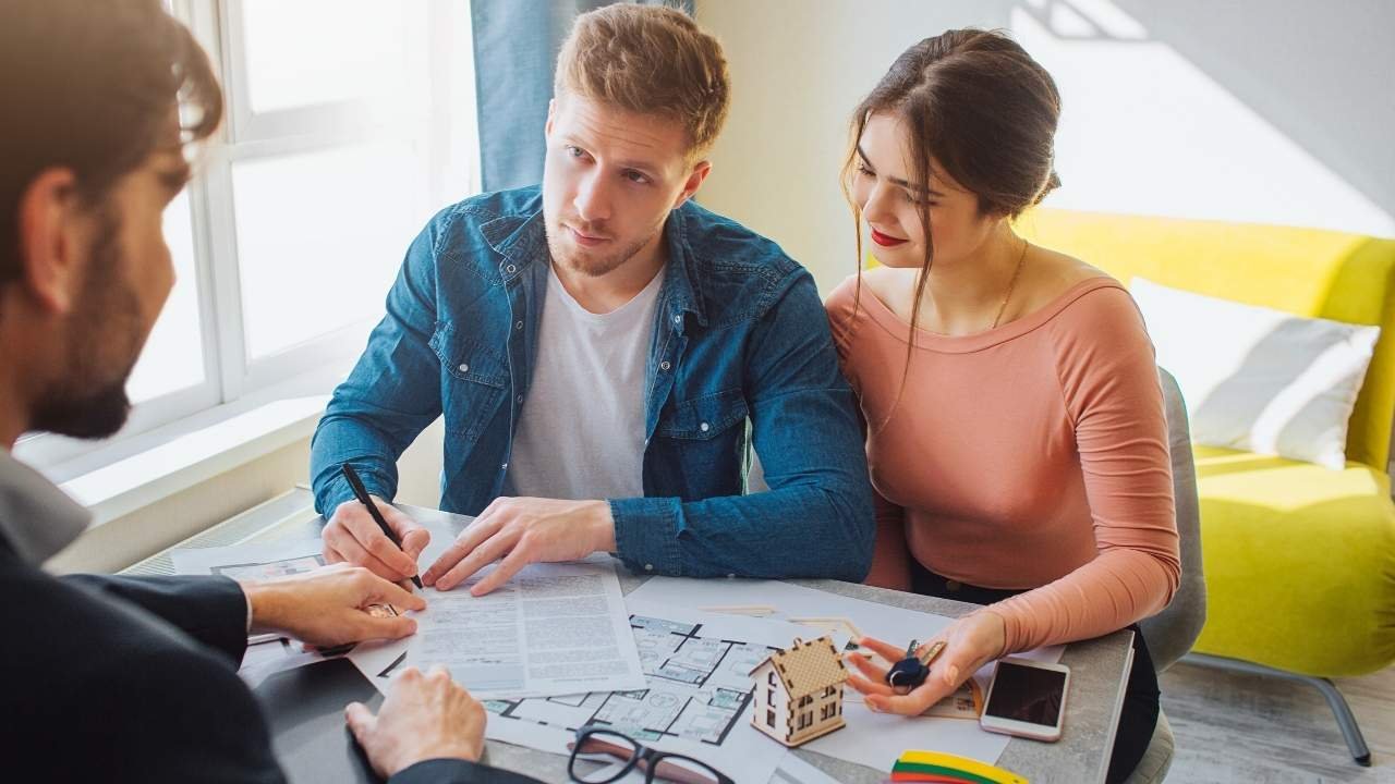 Casal sentado em uma mesa com um corretor, olhando para um contrato, um modelo de casa e uma planta baixa, representando as etapas de como funciona a compra de um apartamento.