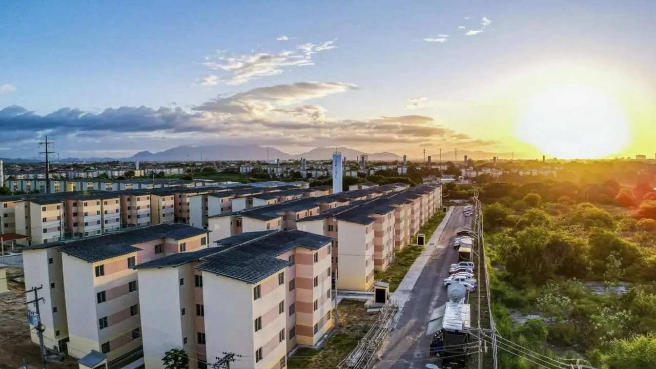 Prédios de apartamentos do programa Minha Casa Minha Vida, em um projeto para a Faixa 4. O sol se põe no horizonte, com montanhas à distância. 