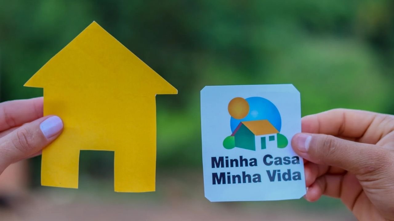 Mãos seguram um recorte de papel em forma de casa amarela e a logomarca do programa habitacional Minha Casa Minha Vida. Este cenário representa o acesso à moradia através do programa, incluindo a Faixa 4. 