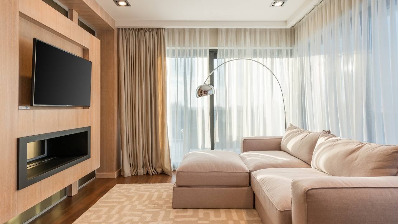Ambiente de sala de TV com sofá grande, cortinas e um painel de madeira claro com uma TV e um nicho, demonstrando uma opção elegante de painel para sala de tv.