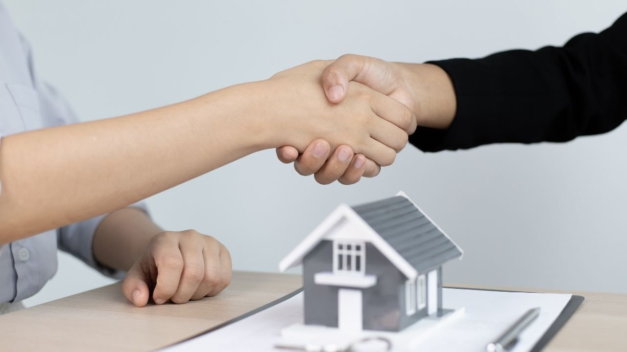 Duas pessoas se cumprimentam com um aperto de mãos, fechando o negócio para a aquisição do seu primeiro imóvel, com uma miniatura de uma casa e as chaves em primeiro plano sobre a mesa.