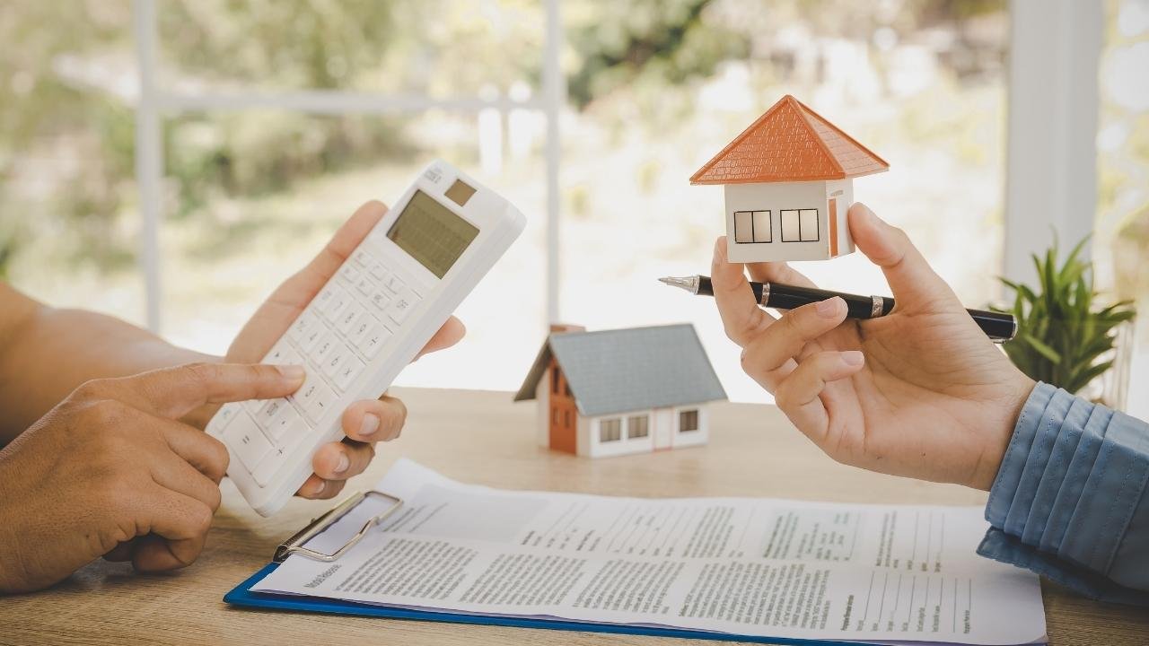 Duas pessoas se preparam para assinar um contrato, uma segurando uma caneta e uma miniatura de casa, enquanto a outra usa uma calculadora. A imagem mostra a fase final do processo de comprar imóvel na planta, destacando a importância da análise financeira e da assinatura de documentos.
