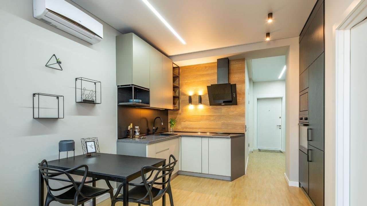 Ambiente de cozinha e sala de jantar integrado em tons neutros, com iluminação para apartamento pequeno. No teto, duas luminárias de trilho com spots e uma luminária linear central destacam diferentes áreas. Na parede sobre a bancada, dois pendentes de design moderno adicionam um toque elegante, enquanto armários em cinza, madeira e preto completam o design.