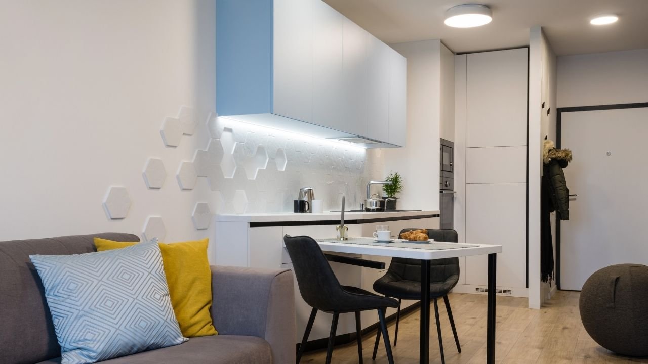 Uma sala de estar e cozinha integradas com iluminação para apartamento pequeno. Uma luminária de teto circular e spots no teto e embaixo dos armários da cozinha proporcionam iluminação funcional e decorativa. A sala tem um sofá cinza com almofadas amarelas e o espaço conta com móveis brancos e um toque de azul claro, mantendo a área clara e espaçosa.