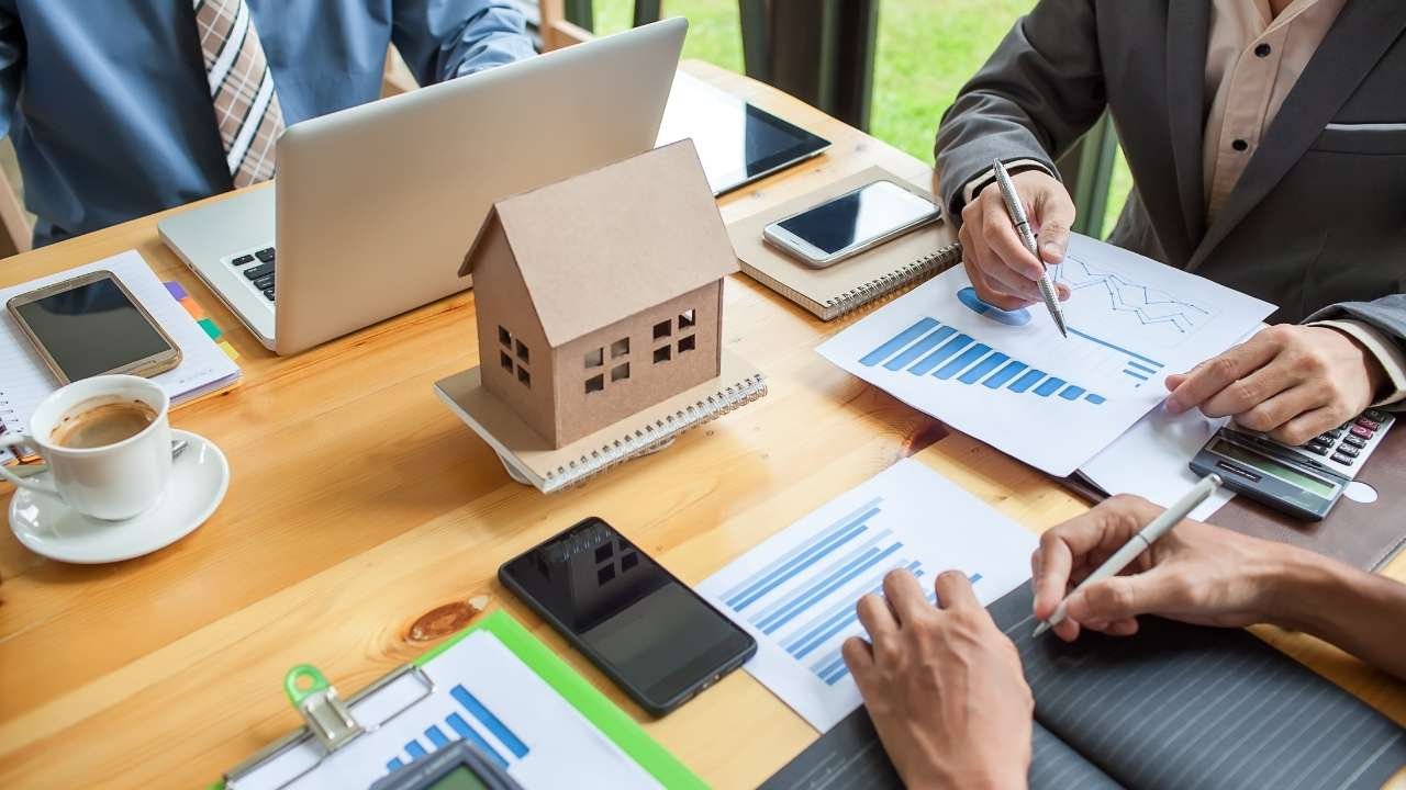 Pessoas sentadas em uma mesa analisando gráficos e documentos. Há uma calculadora, um celular e uma miniatura de casa. A cena reflete o processo de estudo e comparação de diferentes opções de crédito para decidir qual o melhor financiamento imobiliário.