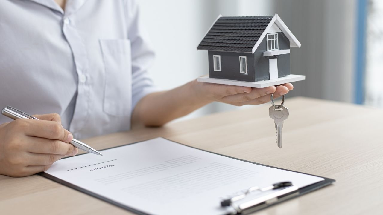 Uma pessoa segura uma miniatura de casa com a chave pendurada, enquanto se prepara para assinar um contrato. A imagem retrata o momento de formalização do negócio, simbolizando a busca por qual o melhor financiamento imobiliário para a compra da casa própria.