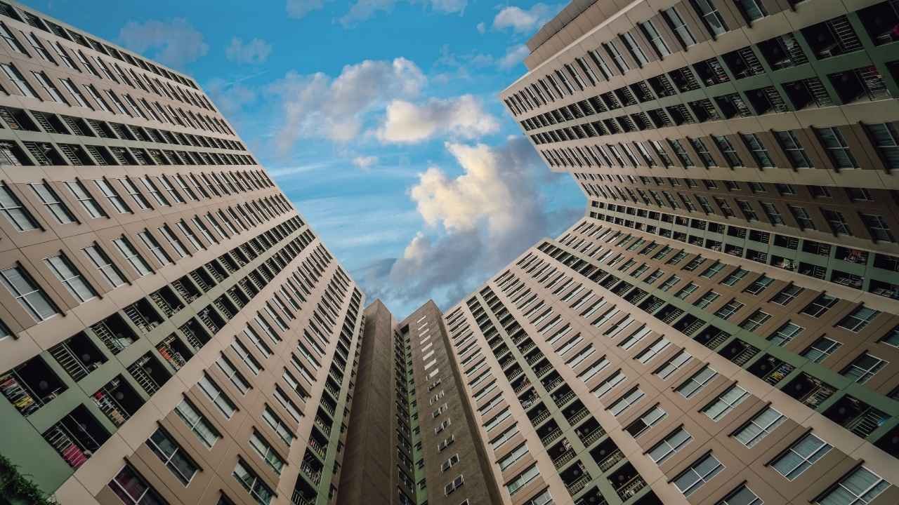 Vista de baixo para cima de três prédios de apartamentos altos e modernos, com fachadas semelhantes. As construções se encontram em um ângulo que enquadra o céu azul com nuvens brancas, ilustrando a dimensão de uma propriedade coletiva e a importância da taxa de condomínio para a manutenção das estruturas.