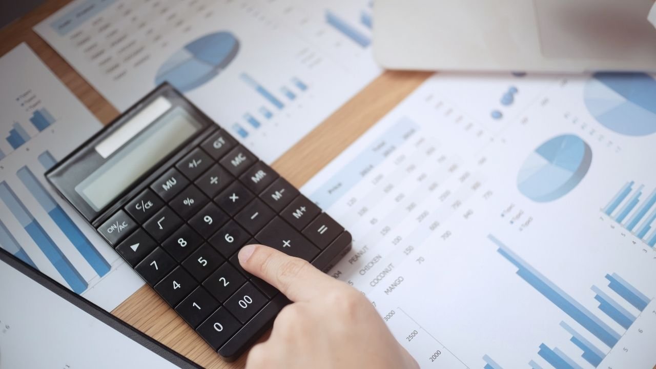 Uma pessoa usando uma calculadora em uma mesa com vários papéis e gráficos de análise financeira. A imagem representa o cálculo e a gestão da taxa de condomínio, mostrando a parte administrativa e financeira envolvida na cobrança e na distribuição dos custos do prédio.
