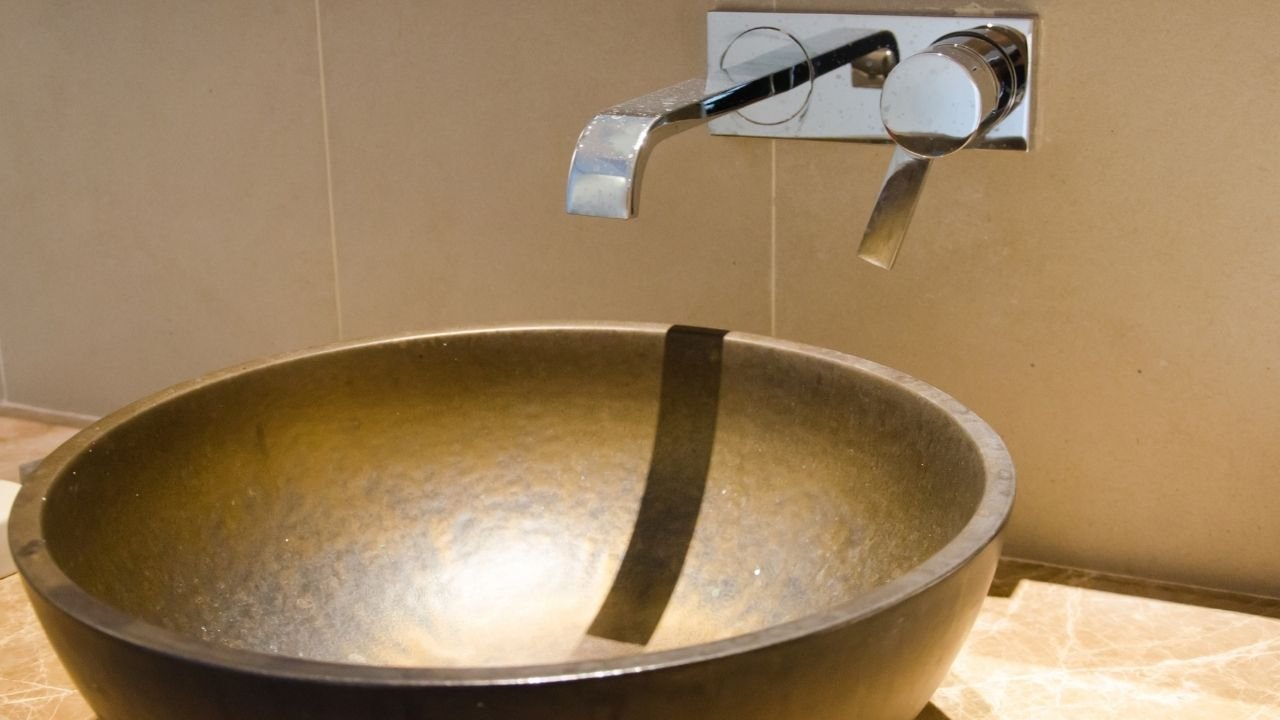 Detalhe de uma cuba de apoio redonda com textura rústica e acabamento escuro, instalada sob uma torneira de parede cromada moderna.