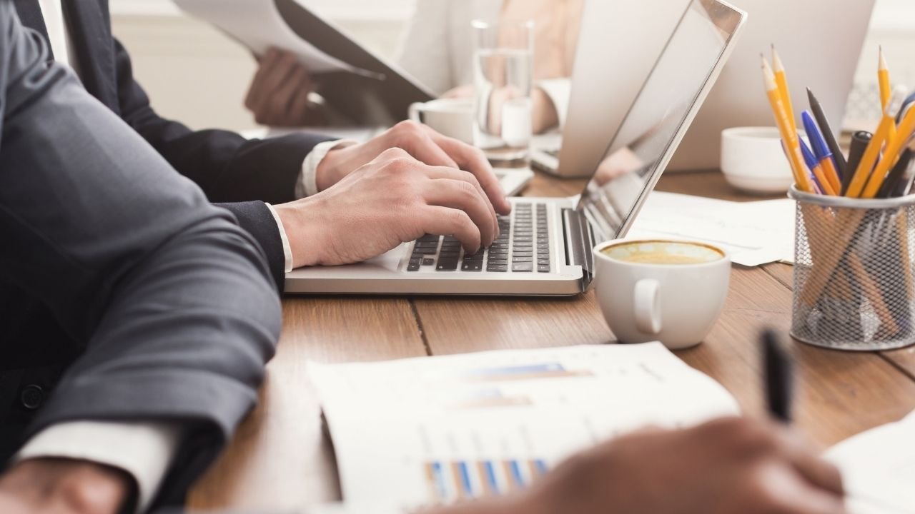 Vista em ângulo de um ambiente de trabalho onde uma pessoa digita em um notebook, com café e materiais de escritório na mesa.