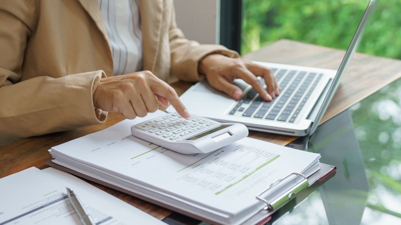 Close das mãos de uma profissional utilizando uma calculadora branca sobre planilhas impressas, com um notebook ao lado.