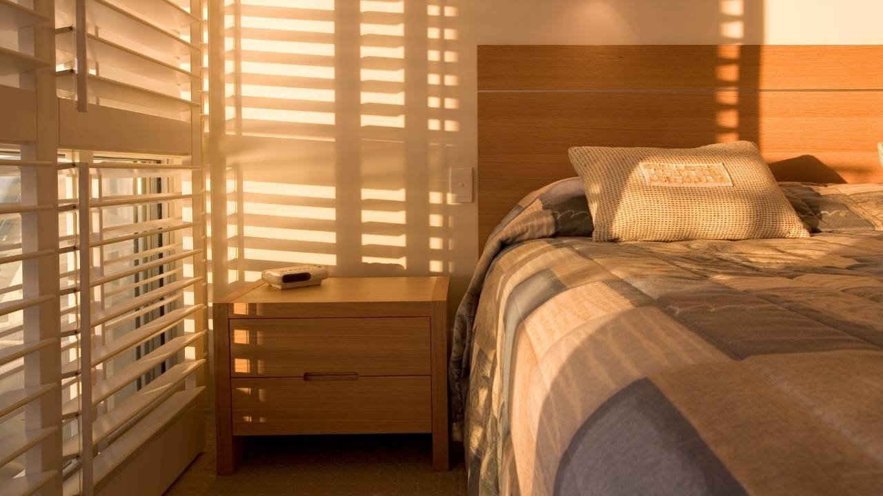 Quarto aconchegante iluminado pelo pôr do sol, destacando um dos tipos de cortinas para quarto mais funcionais: a persiana de madeira com ripas horizontais. A luz atravessa as lâminas criando um padrão geométrico de sombras sobre a mesa de cabeceira de madeira e a cama arrumada.