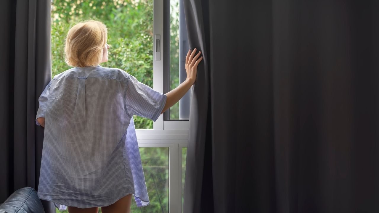 Uma mulher de costas abre uma cortina cinza escura em um quarto iluminado. A imagem demonstra a eficiência da cortina blackout, um dos principais tipos de cortinas para quarto para quem busca total controle da luminosidade externa e privacidade.