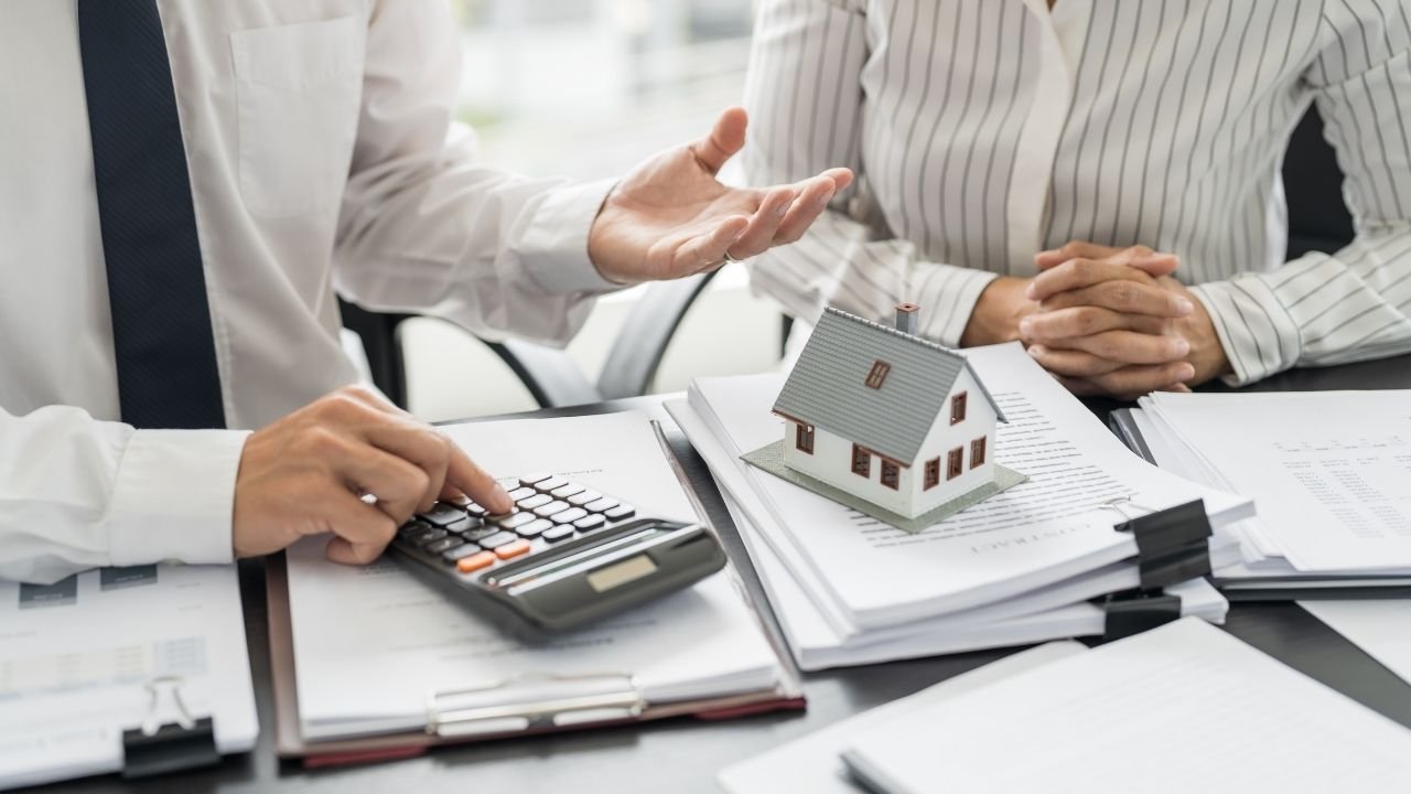 Consultor imobiliário usando uma calculadora e gesticulando para um cliente durante reunião sobre documentos e uma maquete de casa.