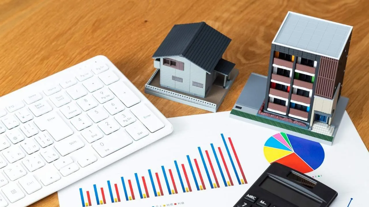 Uma mesa de escritório com teclado, calculadora e miniaturas de uma casa e um prédio sobre gráficos financeiros de barras e pizza.