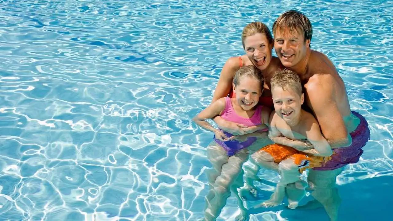 Uma família feliz com pai, mãe e dois filhos sorrindo dentro de uma piscina num dia ensolarado.