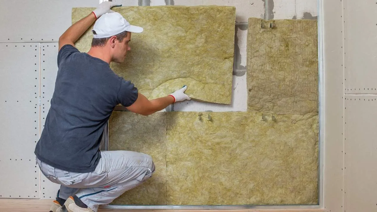 Profissional da construção civil instalando placas rígidas de lã de rocha para isolamento térmico em uma parede interna.